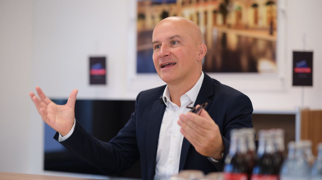 Ein Mann im Anzug macht eine Geste mit der Hand, während er in einem Büro spricht. Er hat eine Brille in der Hand und scheint an einem Gespräch beteiligt zu sein.