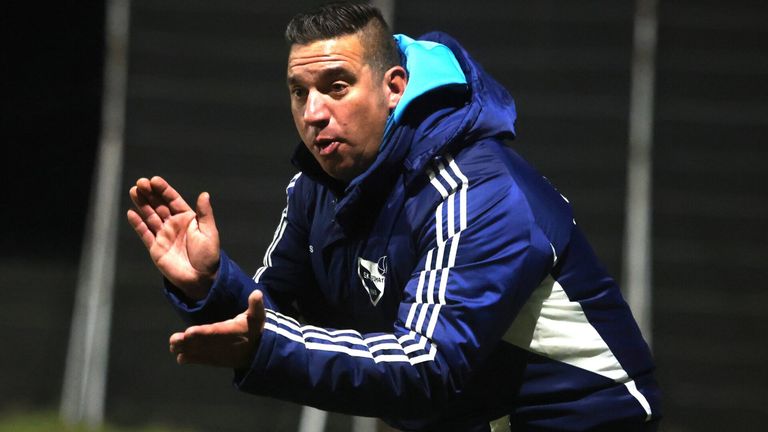 Ein Mann in einem blauen Adidas-Jacke gestikuliert und schaut nach rechts. Die Jacke hat ein weißes Logo auf der linken Brust.