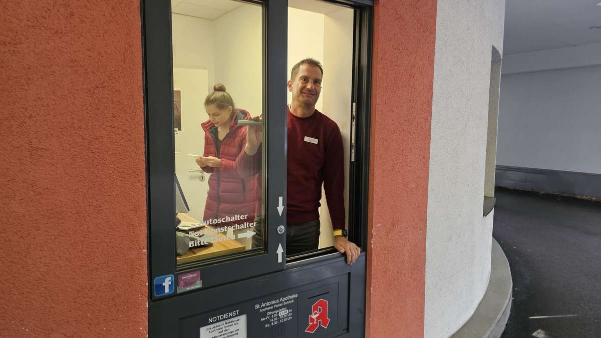 A man stands outside a door, smiling, with a woman behind him inside the room. The door has a sign that reads 'St. Antonius Apotheke' and a red 'A'. The woman is holding a card and appears to be texting.