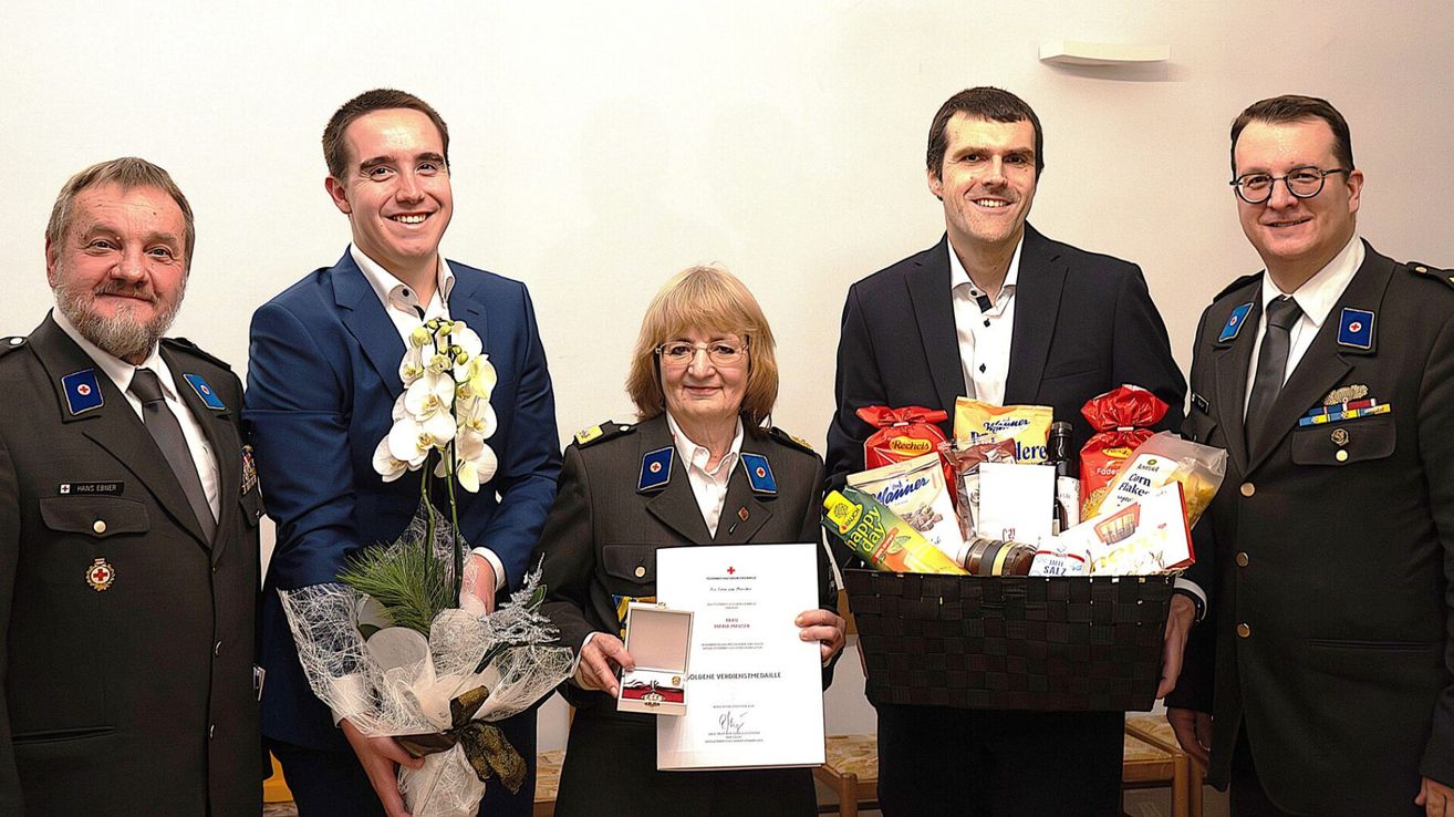 Three individuals stand together smiling, one holding a bouquet, another holding a certificate and medal, and the third holding a basket of snacks.