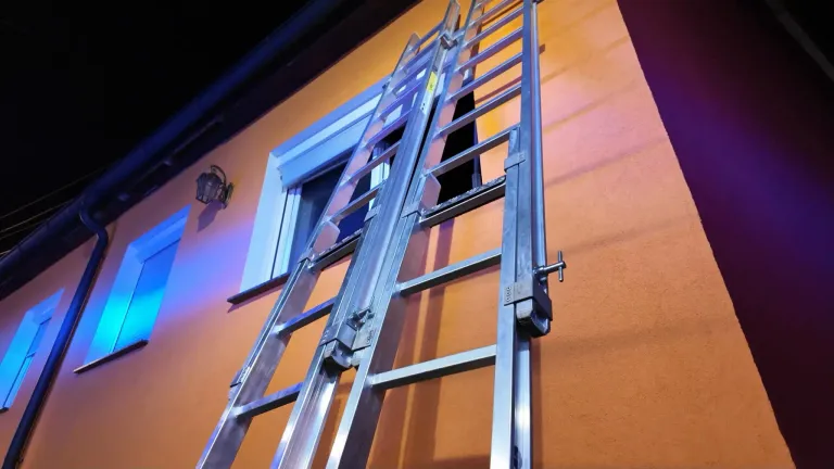 Eine silberne Leiter ist an einem orangefarbenen Gebäude mit einem Fenster positioniert und wirft einen Schatten an die Wand.