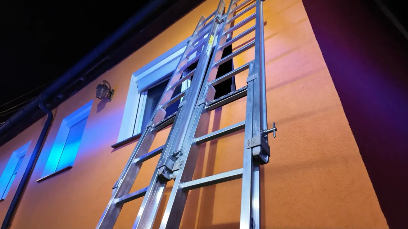 Eine silberne Leiter ist an einem orangefarbenen Gebäude mit einem Fenster positioniert und wirft einen Schatten an die Wand.