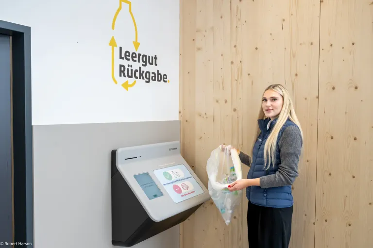 Eine Frau in einer blauen Weste hält einen Plastikbeutel vor einem Gerät. Das Gerät ist mit 'Leergut Rückgabe' beschriftet. Hinter ihr zeigt eine Holzwand das gleiche Logo.