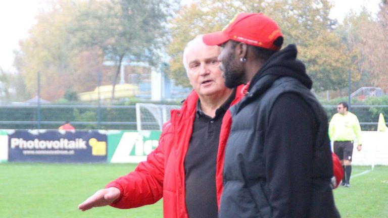 Zwei Männer stehen auf einem Outdoor-Gelände, wahrscheinlich auf einem Fußballfeld. Der Mann auf der linken Seite zeigt auf etwas, und der Mann auf der rechten Seite schaut ihn an. Sie tragen beide Jacken und Mützen. Hinter ihnen ist ein unscharfer Hintergrund von einem Zaun, Bäumen und einem Gebäude.