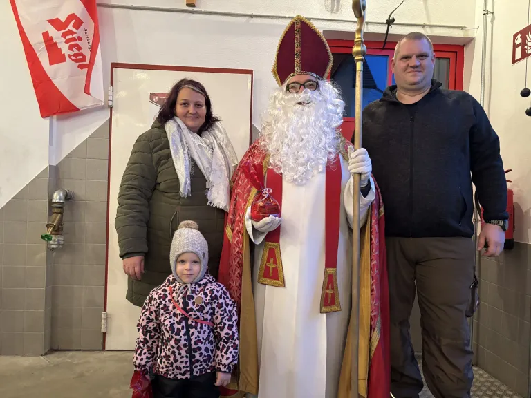 Eine Familie mit einem kleinen Kind steht neben einem Mann in einer Weihnachtsmann-Verkleidung und posiert für ein Foto. Der Weihnachtsmann hält einen goldenen Stab, und sie sind drinnen.