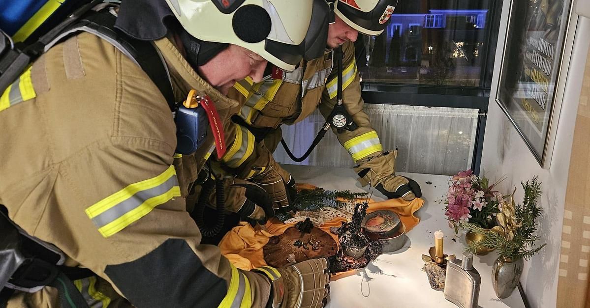 Zwei Feuerwehrleute in Schutzkleidung arbeiten an einem Gegenstand auf einem Tisch mit Blumen und Kerzen. Sie beugen sich über einen beschädigten Gegenstand, möglicherweise während einer Trainingsübung.
