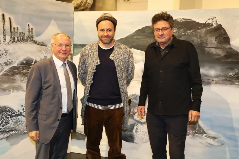 Drei Männer stehen vor einer großen Wandmalerei. Der Mann auf der linken Seite trägt einen Anzug, der Mann in der Mitte eine Strickjacke und der Mann auf der rechten Seite ein schwarzes Hemd. Sie lächeln.