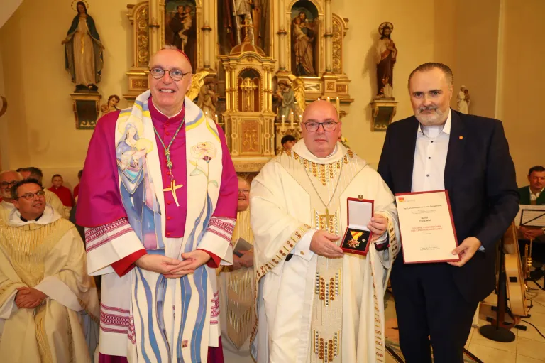 Drei Männer in formeller Kleidung stehen in einer Kirche. Einer hält einen Preis. Hinter ihnen steht ein Priester mit einem Kreuz. Hinter dem Priester befindet sich ein Altar mit Statuen.