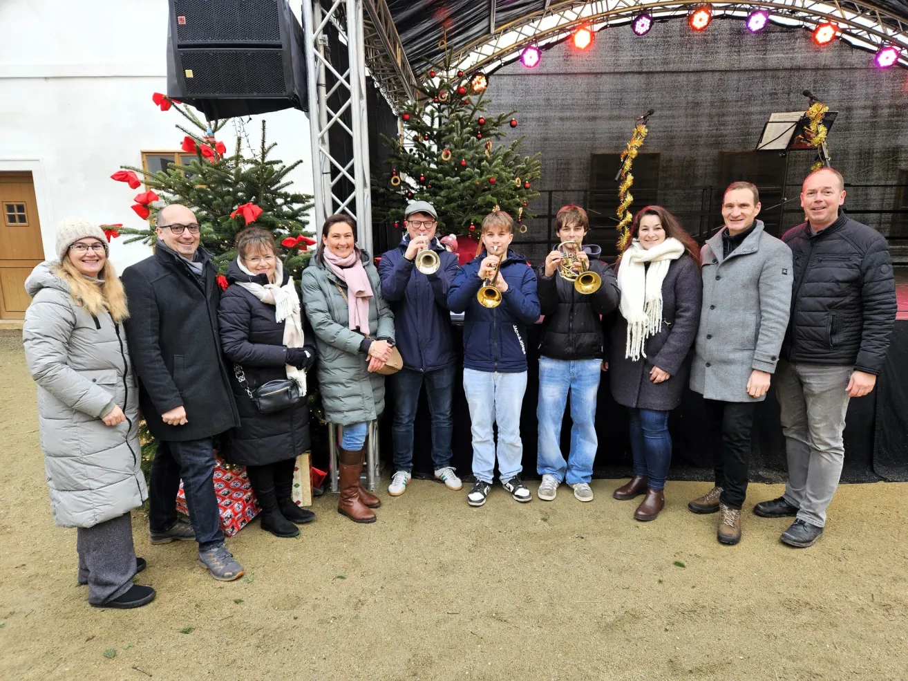 A group of people, both men and women, are standing in front of a decorated Christmas tree, holding musical instruments, possibly trumpets. They are smiling and seem to be posing for a photo. Behind them is a stage with a speaker and a microphone.