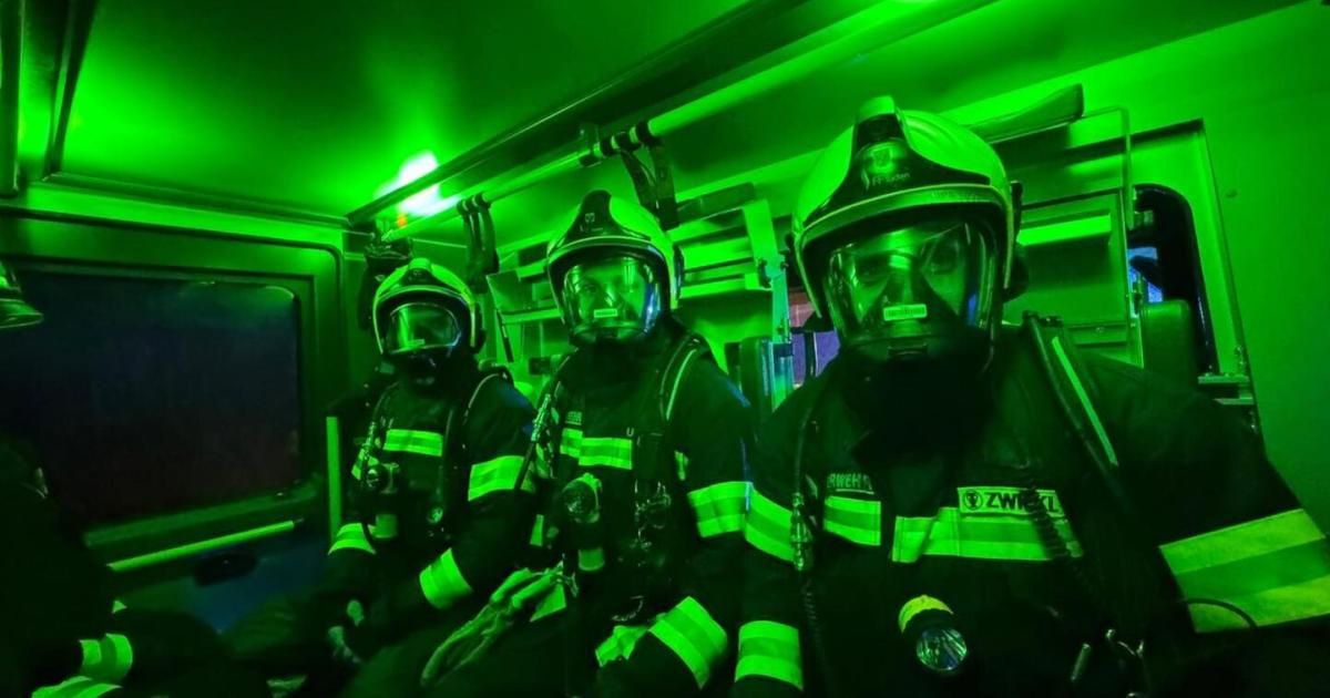 Drei Feuerwehrleute in voller Ausrüstung sitzen in einem von grünem Licht erleuchteten Fahrzeug. Jeder trägt einen Helm mit Maske und Handschuhen. Sie scheinen einsatzbereit zu sein.