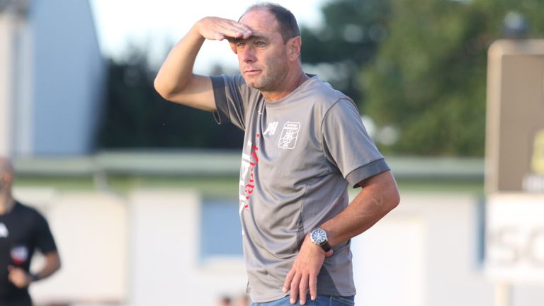 Ein Mann in einem grauen Shirt mit dem Wort 'PH' und einer Uhr steht draußen und schattet seine Augen mit der Hand.