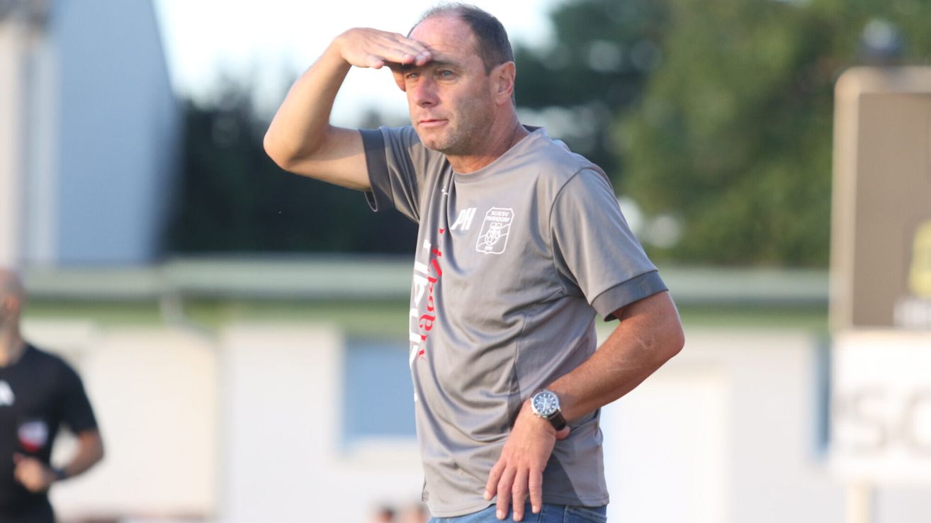 Ein Mann in einem grauen Shirt mit dem Wort 'PH' und einer Uhr steht draußen und schattet seine Augen mit der Hand.