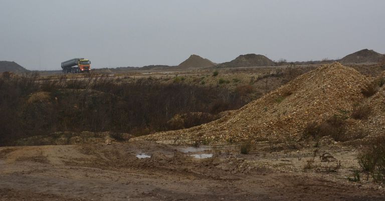 Ein gelber Lastwagen fährt auf einer unbefestigten Straße mit einem Haufen Steine und Erde im Hintergrund. Der Himmel ist bedeckt.