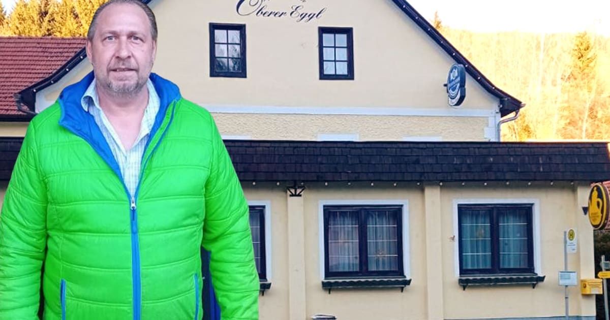 A man in a green jacket is standing in front of a building with the sign 'Oberer Egel'. The building has a black roof, glass windows, and a small awning.