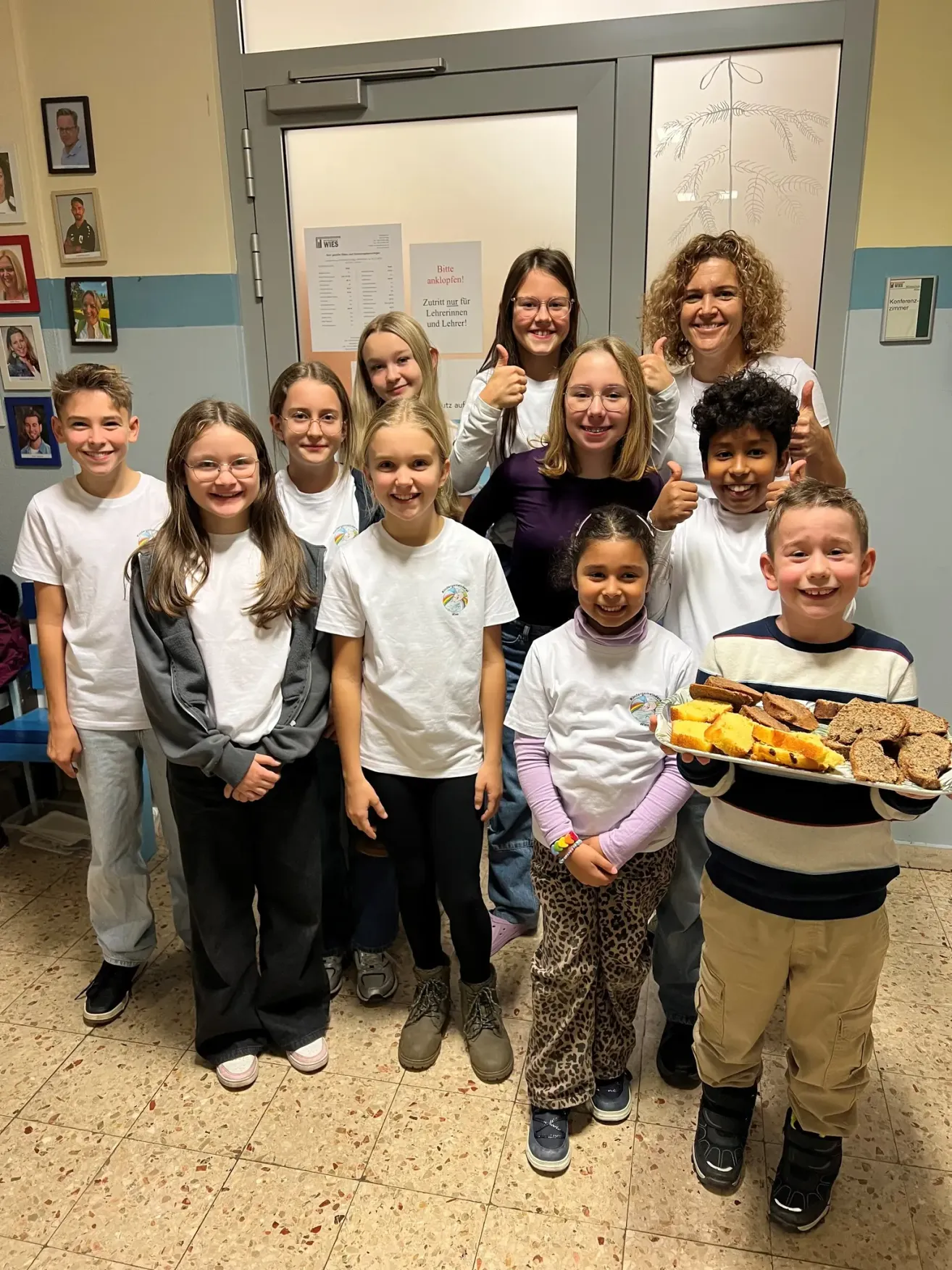Eine Gruppe von Kindern und zwei Frauen steht in einem Klassenzimmer. Ein Kind hält einen Teller mit Essen. Sie lächeln und zeigen Daumen hoch.