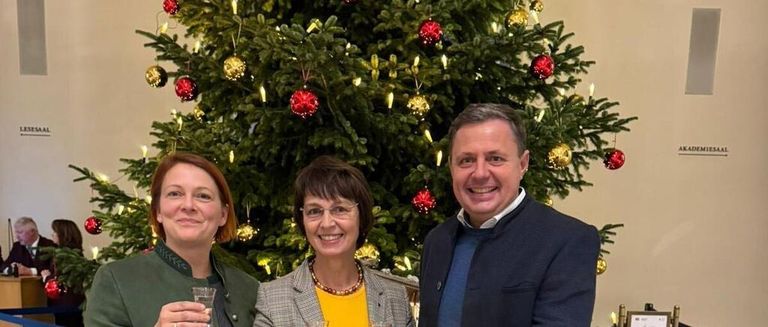 Three people, two women and a man, are standing together in front of a Christmas tree, smiling and holding glasses of wine.