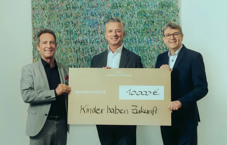 Three men hold a large check with the words 'Children have a future' and '10,000€'. They stand in front of a colorful abstract painting.