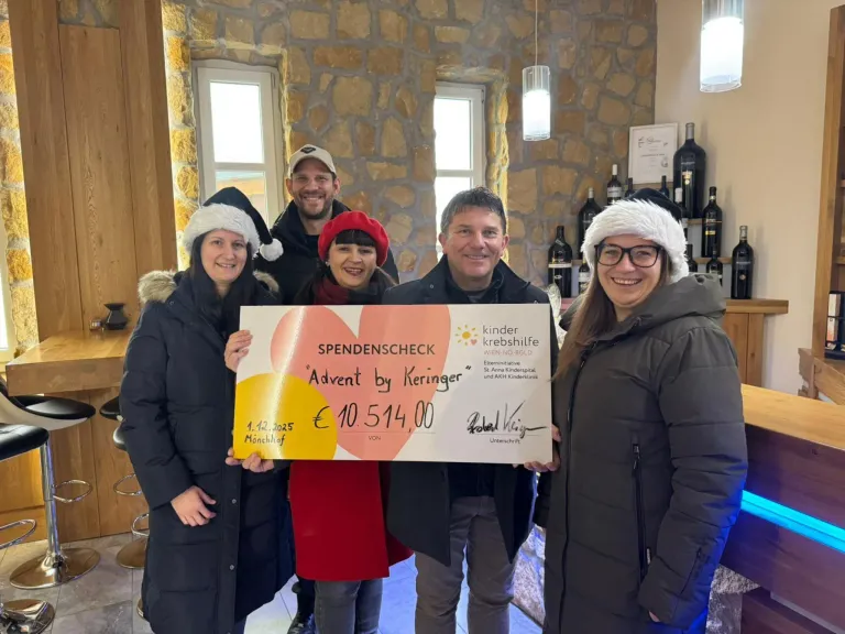 Fünf Erwachsene stehen in einem Raum und lächeln. Sie halten einen großen Scheck über 10.514,00 Euro, ausgestellt von Keringer, datiert auf den 1. Dezember 2025. Die Frau in der Mitte trägt einen roten Hut. Dahinter befinden sich Regale mit Weinflaschen.