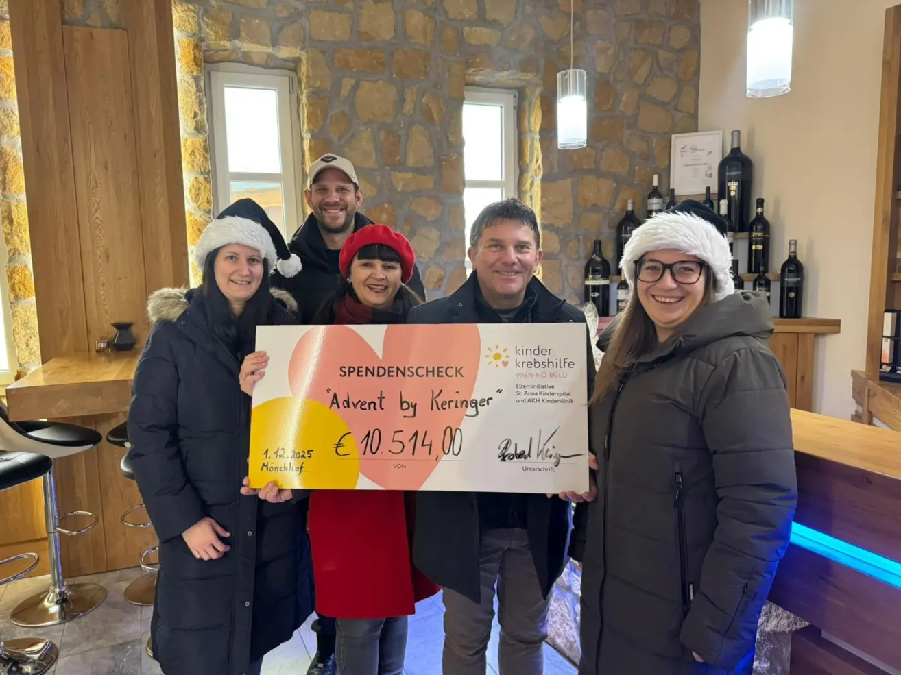 Fünf Erwachsene stehen in einem Raum und lächeln. Sie halten einen großen Scheck über 10.514,00 Euro, ausgestellt von Keringer, datiert auf den 1. Dezember 2025. Die Frau in der Mitte trägt einen roten Hut. Dahinter befinden sich Regale mit Weinflaschen.