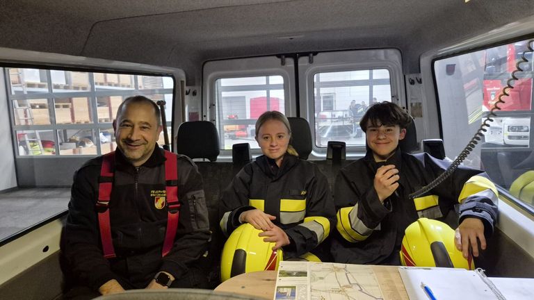 Drei Personen in Feuerwehruniformen sitzen in einem Fahrzeug. Ein Mann links lächelt, eine Frau in der Mitte hält einen gelben Helm, und ein Junge rechts posiert für das Foto. Vor dem Fahrzeug steht ein Gebäude mit einem roten Tank.