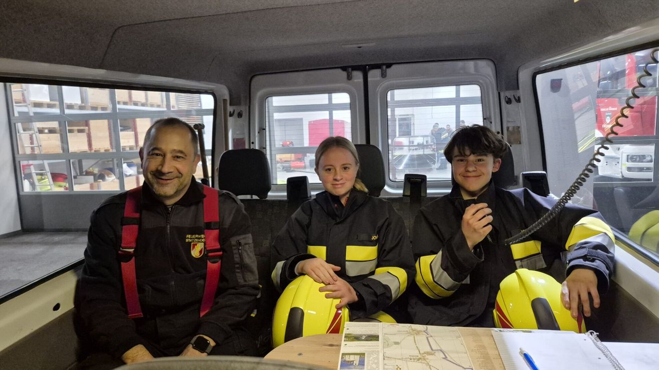 Drei Personen in Feuerwehruniformen sitzen in einem Fahrzeug. Ein Mann links lächelt, eine Frau in der Mitte hält einen gelben Helm, und ein Junge rechts posiert für das Foto. Vor dem Fahrzeug steht ein Gebäude mit einem roten Tank.