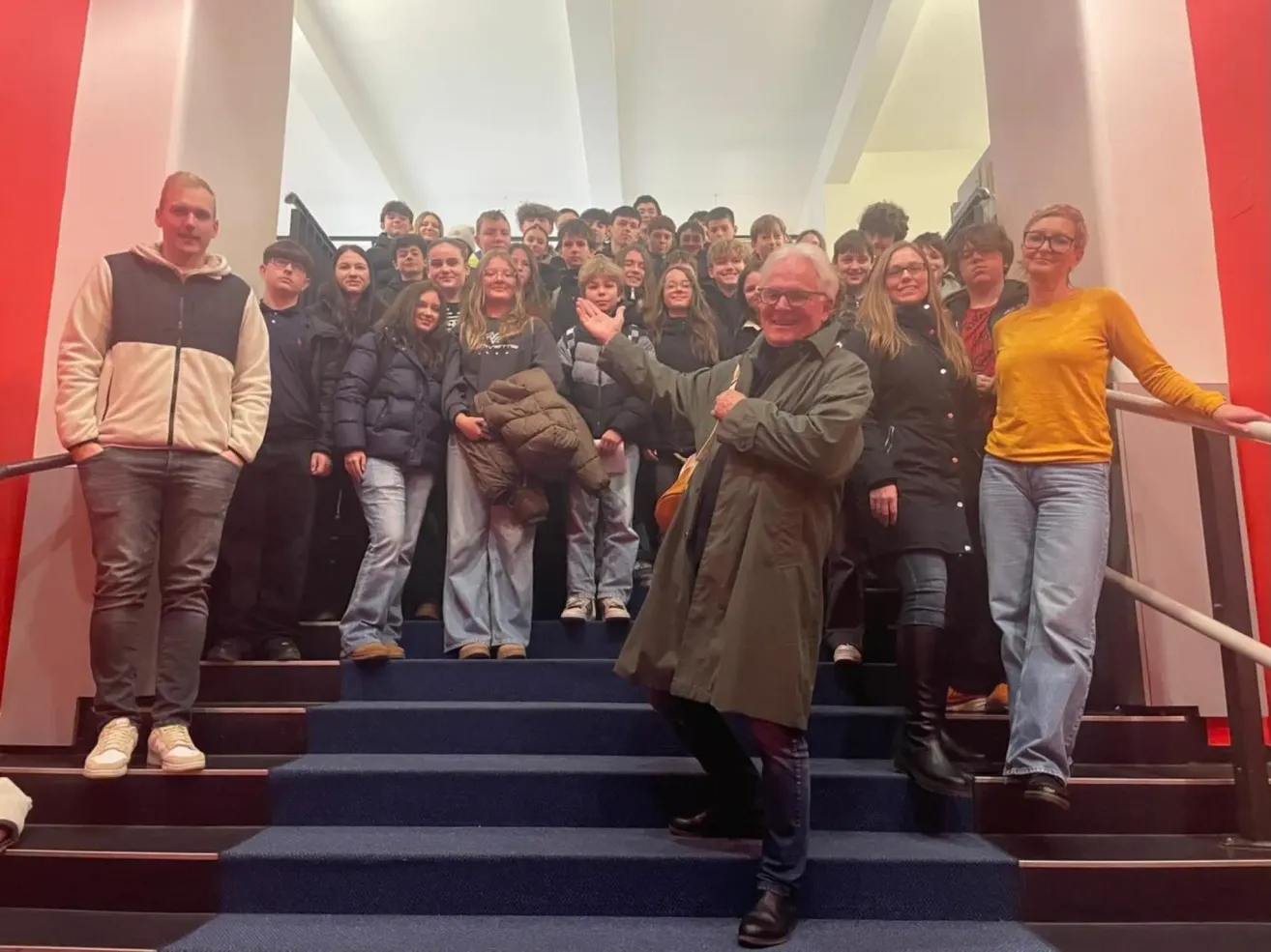 Eine Gruppe von Menschen, darunter ein Mann in einem grünen Mantel, steht auf blauen Teppichstufen. Sie scheinen für ein Foto zu posieren.