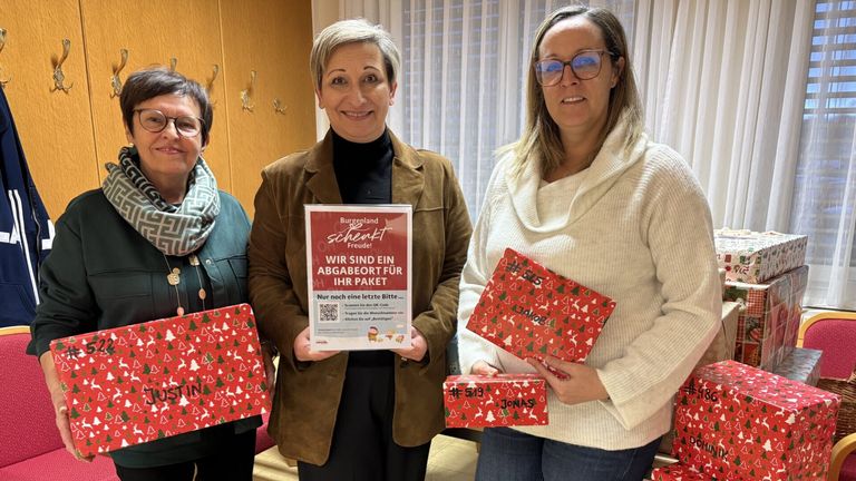 Drei Frauen halten rote Geschenkboxen mit dem Text 'Burgenland sendet Freude!'. Die Frau in der Mitte hält ein Schild mit einem QR-Code. Sie lächeln für die Kamera.