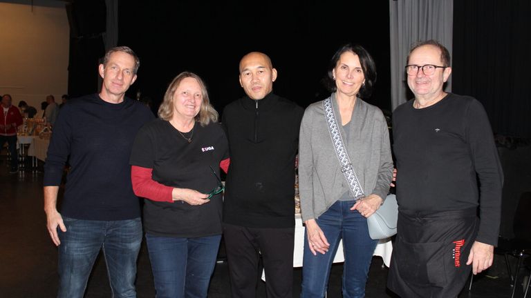 Five people pose for a photograph. Three women and two men, all smiling, stand close together. The woman on the left holds a pair of glasses. Behind them, a table with various items is visible.