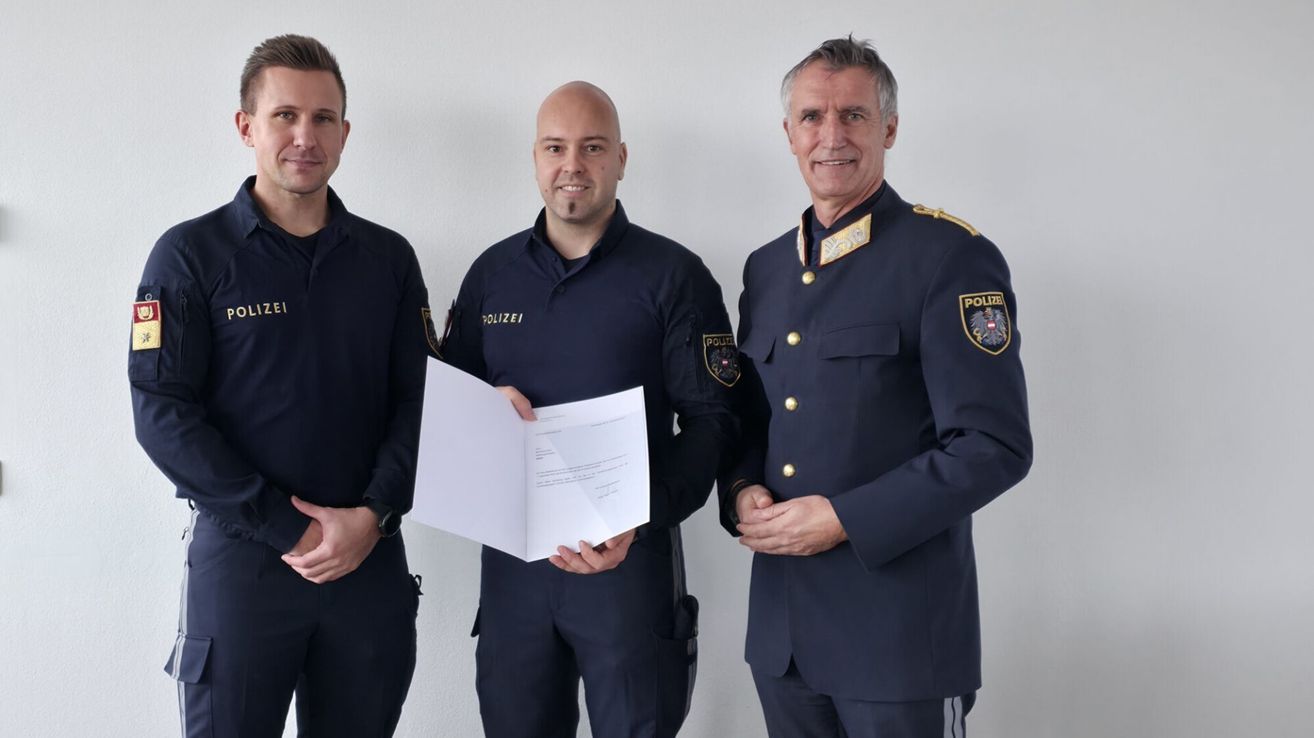 Drei Männer in Uniform, zwei in Marineblau und einer in höherem Rang, posieren mit einem Dokument, wahrscheinlich eine Auszeichnung. Sie stehen vor einem schlichten weißen Hintergrund.