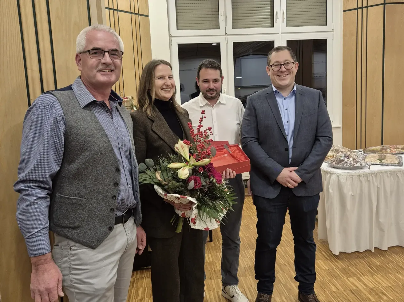 Vier Menschen lächeln in einem Raum, zwei Männer und zwei Frauen. Die Frau in der Mitte hält einen Blumenstrauß und eine rote Schachtel. Hinter ihnen ist eine Glastür und ein Tisch mit weißer Tischdecke.