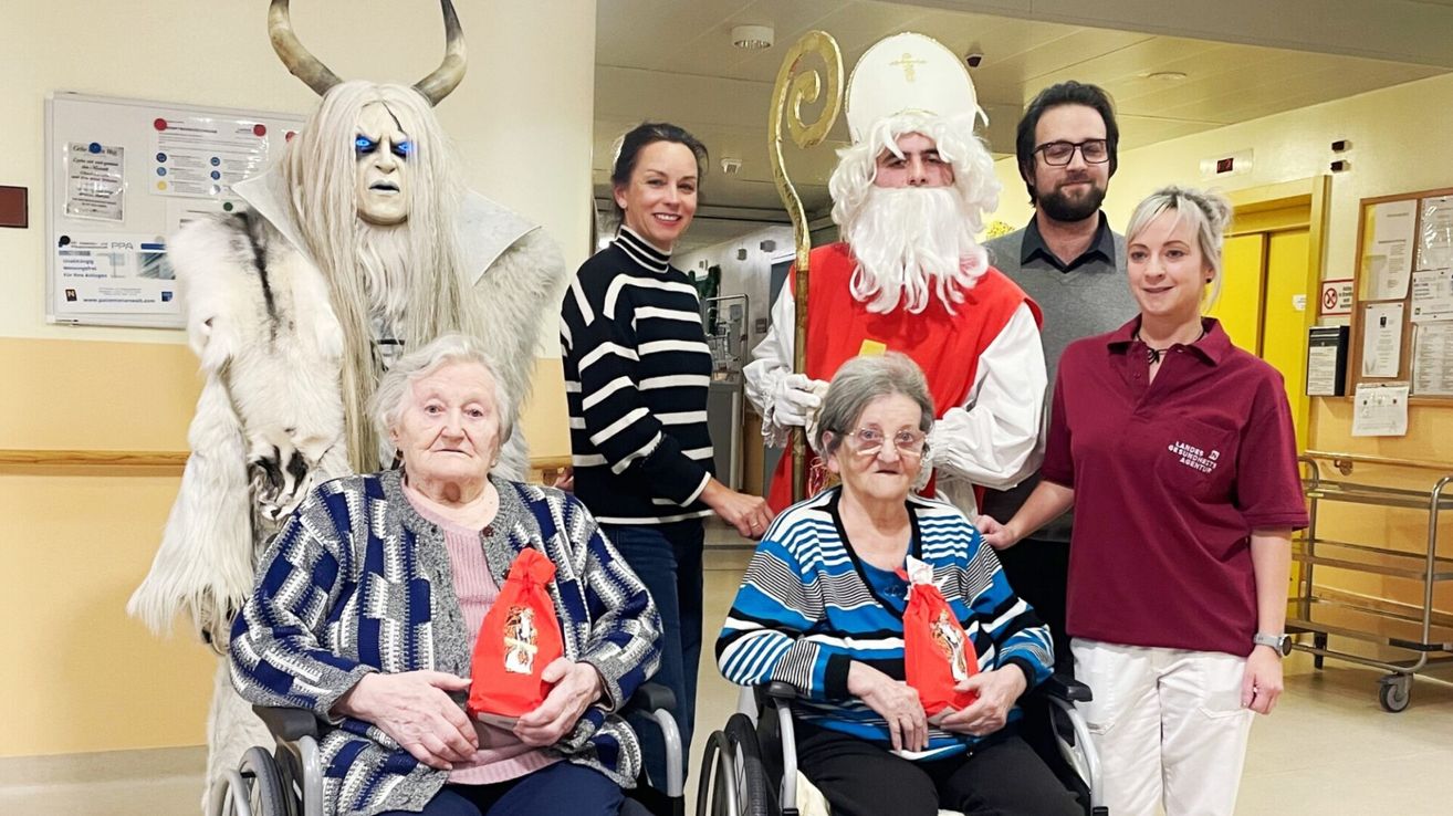 Two elderly women in wheelchairs are receiving gifts from a Santa Claus and Krampus. A woman stands behind them smiling. Behind her, a man in a red costume holds a staff.