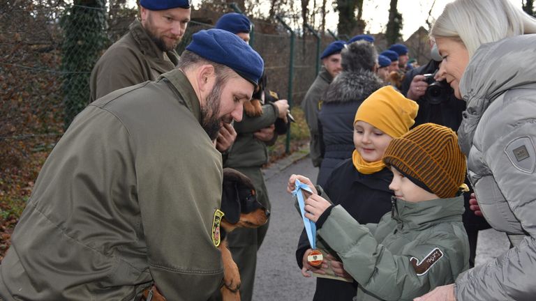 Ein Mann in Uniform hält einen Hund, während ein junges Mädchen eine Medaille von einer anderen Person, möglicherweise einem Trainer, erhält, während andere zusehen.