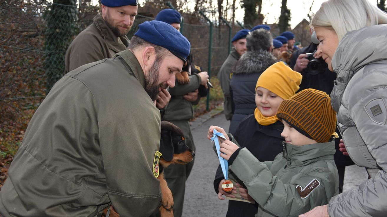 Ein Mann in Uniform hält einen Hund, während ein junges Mädchen eine Medaille von einer anderen Person, möglicherweise einem Trainer, erhält, während andere zusehen.