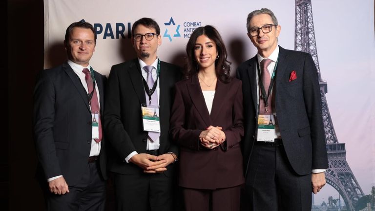 Four individuals, two men and two women, are posing for a photo in front of a backdrop with the text 'COMBAT ANTISEMITISM'. The woman is wearing a brown suit and a necklace. The man on the left is wearing glasses and a tie.