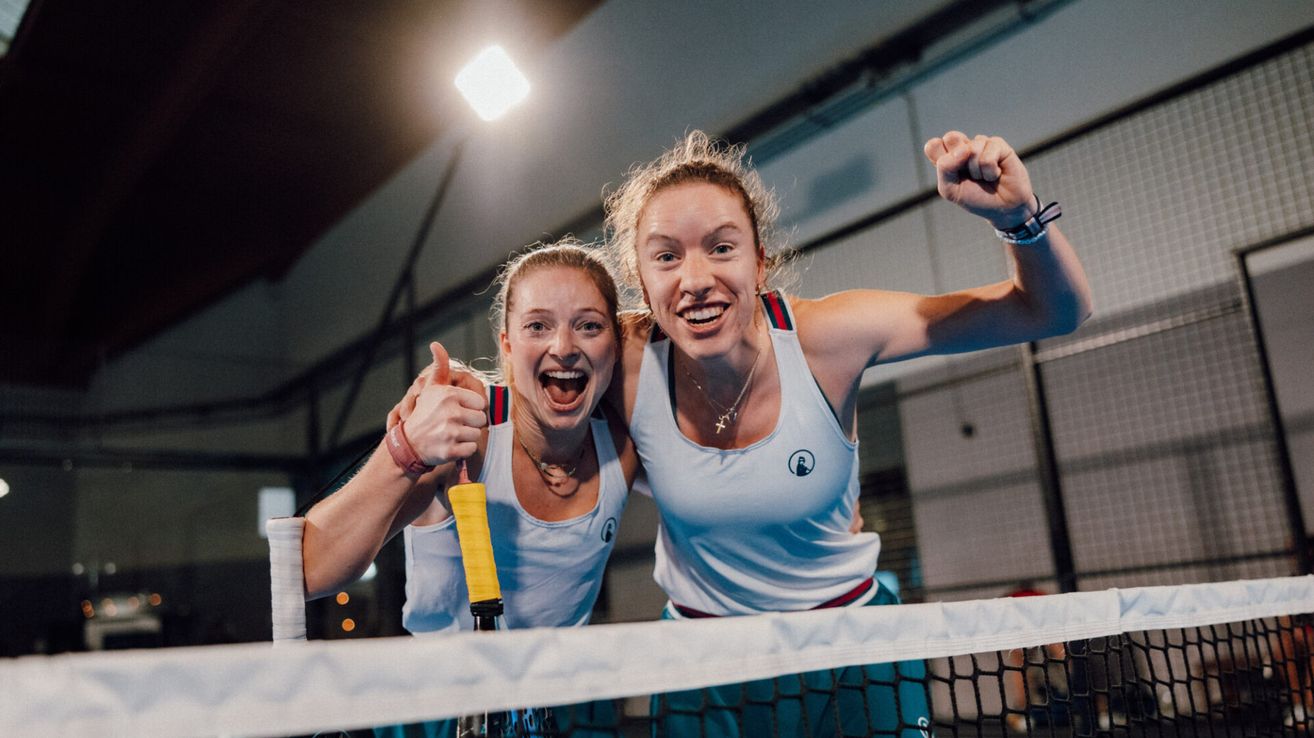 Zwei Frauen in weißen Tanktops und grünen Armbändern stehen hinter einem Tennisnetz, feiern mit erhobenen Daumen und breiten Lächeln, eine hält einen Tennisschläger.