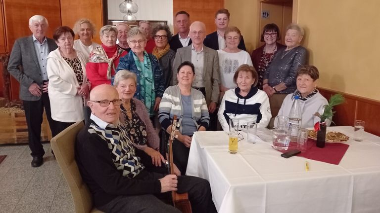 A group of elderly people and some younger individuals gather in a room, posing for a photograph. Some are seated, others stand, with a table in front featuring drinks and glasses.