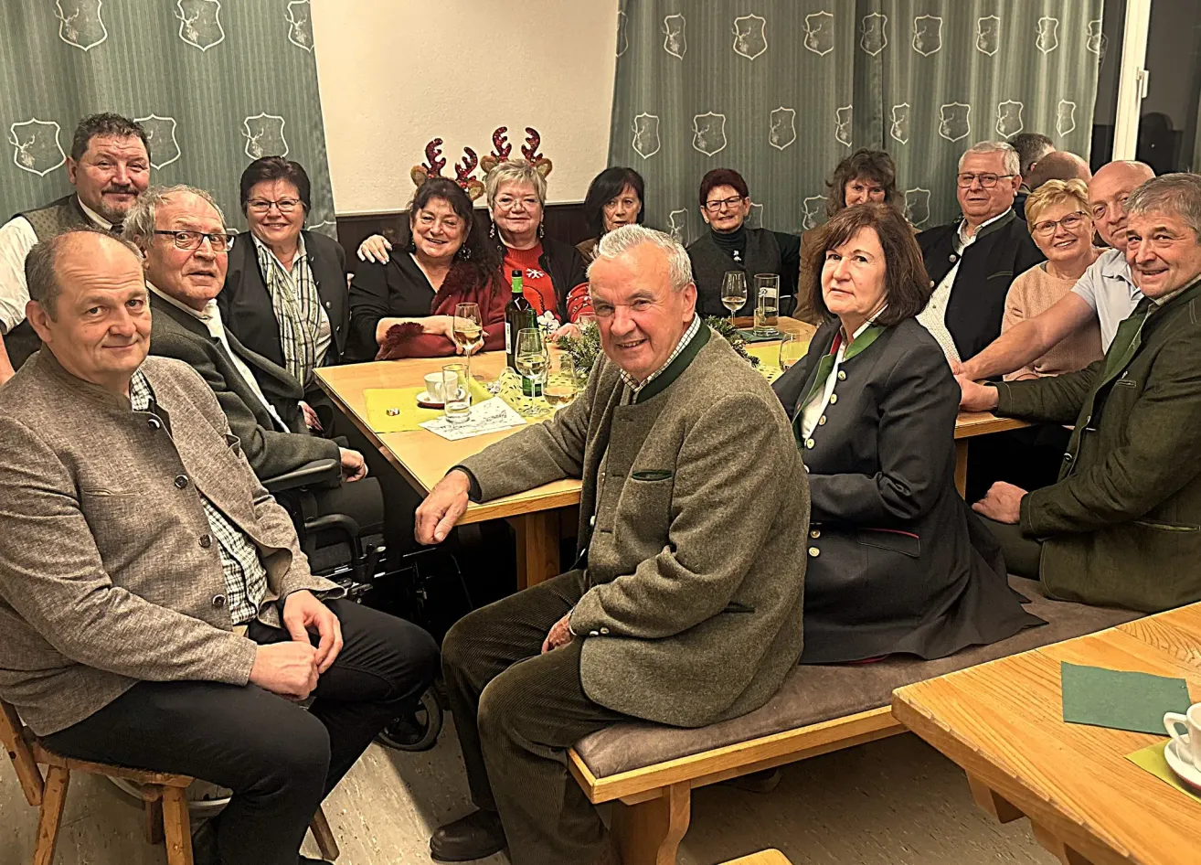 A group of adults are seated around a table, smiling for a photo. They are dressed in formal attire, with some wearing Christmas headbands. The table is set with wine glasses, a bottle, and a vase with greenery.