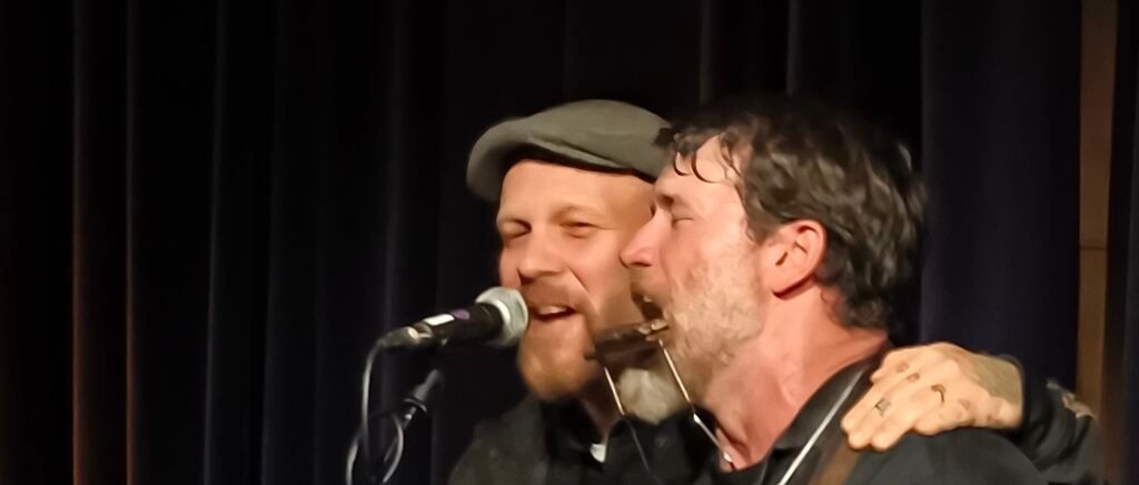 Two men perform on stage, one with a beard singing into a microphone, the other playing a harmonica, with a dark background.