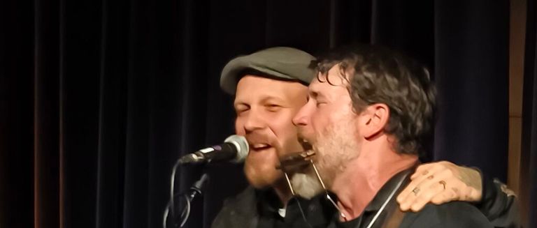 Two men perform on stage, one with a beard singing into a microphone, the other playing a harmonica, with a dark background.