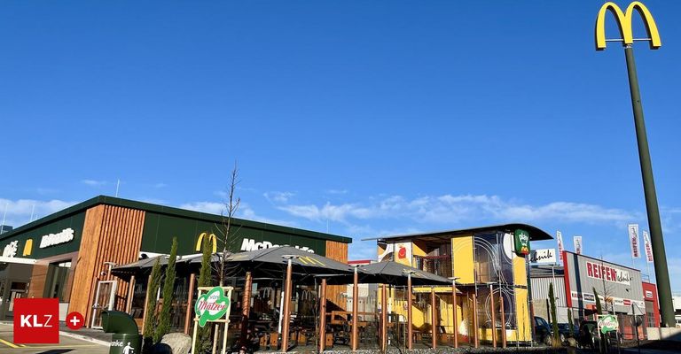 A McDonald's restaurant with a play area beside it, featuring a yellow structure with colorful designs. The area is surrounded by trees and tables under umbrellas.