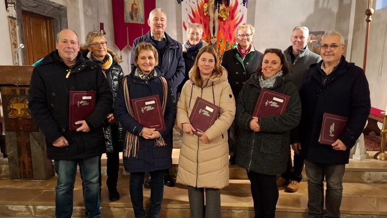 Eine Gruppe von Personen in Wintermänteln steht auf Kirchenstufen, lächelt und hält Bücher. Dahinter ist ein religiöses Banner und ein flammenartiges Gemälde zu sehen.