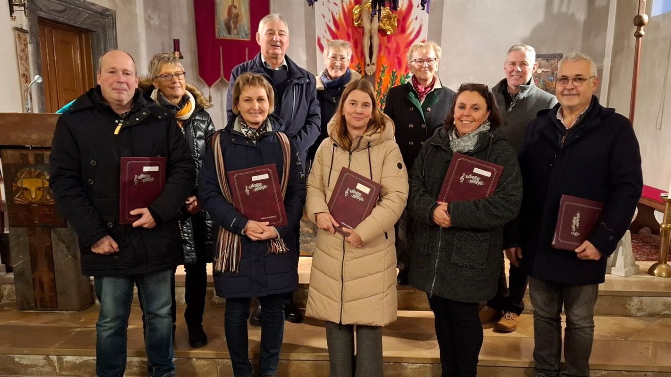 Eine Gruppe von Personen in Wintermänteln steht auf Kirchenstufen, lächelt und hält Bücher. Dahinter ist ein religiöses Banner und ein flammenartiges Gemälde zu sehen.
