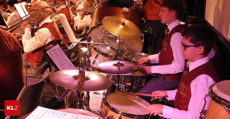 Eine Gruppe von Musikern in roten Westen und Brillen spielt auf der Bühne Trommeln und Becken. Einige lesen Noten, während andere Blechblasinstrumente spielen.