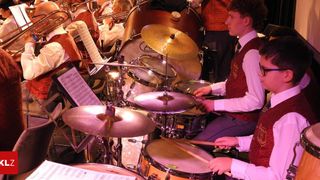 A group of musicians in red vests and glasses are playing drums and cymbals on stage. Some are reading music while others play brass instruments.