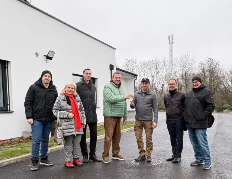 Sechs Personen in Winterkleidung stehen in einer Reihe vor einem Gebäude. Sie scheinen für ein Foto zu posieren. Hinter ihnen ist ein Gebäude mit einem Schornstein zu sehen.