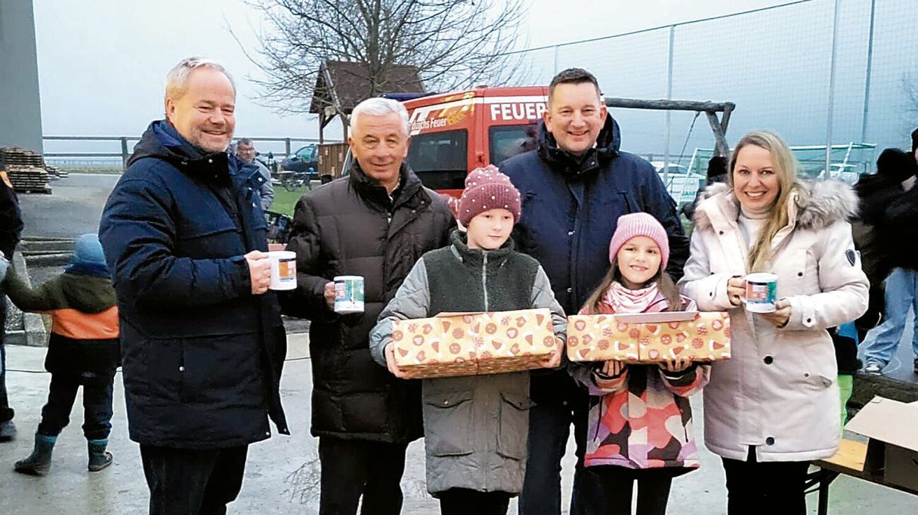 Eine Gruppe von Erwachsenen und Kindern posiert für ein Foto und hält verpackte Geschenke. Dahinter ist ein Feuerwehrauto geparkt, und ein Zaun ist sichtbar. Bäume und ein Haus sind im Hintergrund.