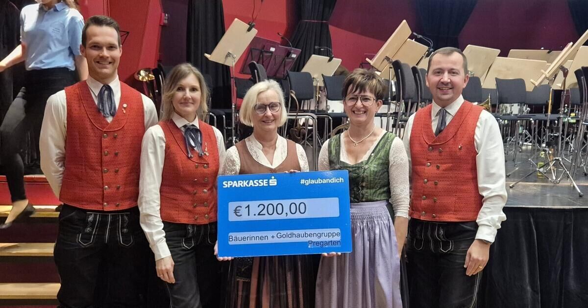 Five people in traditional clothing stand together holding a large blue check. They are standing in front of an orchestra.