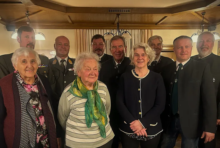 A group of individuals, both men and women, are gathered in a room. They are wearing formal attire. The woman on the left is wearing a scarf. The woman in the middle is wearing a necklace. The man on the right is wearing a tie. The room has a wooden ceiling and a curtain.