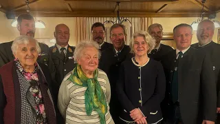 A group of individuals, both men and women, are gathered in a room. They are wearing formal attire. The woman on the left is wearing a scarf. The woman in the middle is wearing a necklace. The man on the right is wearing a tie. The room has a wooden ceiling and a curtain.