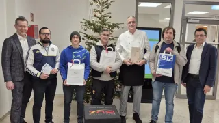 Five men stand in a room next to a Christmas tree and a box with the words Know Your Score Challenge 2024. They all hold bags with the logo Bizz. The man in the middle holds a piece of paper.